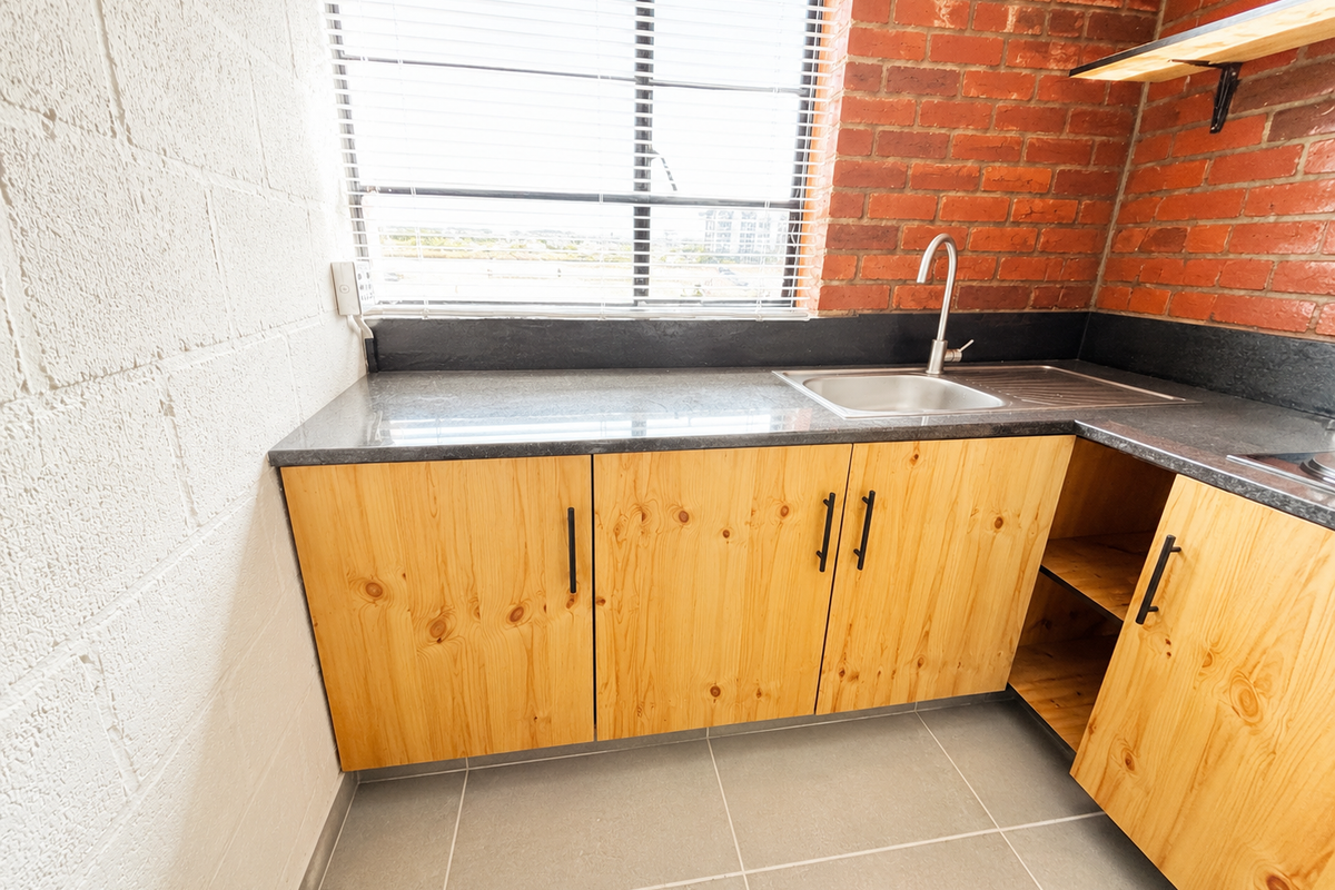 A compact kitchen area featuring exposed brick walls, light wood cabinetry, stone countertops, and a stainless steel sink with a modern faucet.