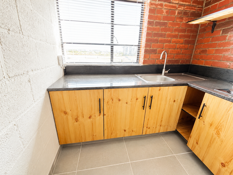 A compact kitchen area featuring exposed brick walls, light wood cabinetry, stone countertops, and a stainless steel sink with a modern faucet.