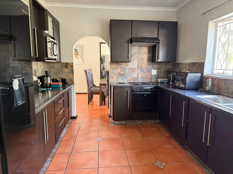 A functional kitchen featuring dark wood cabinetry, tiled splashbacks, built-in oven and hob, and ceramic floor tiles, with an arched opening to the dining area.
