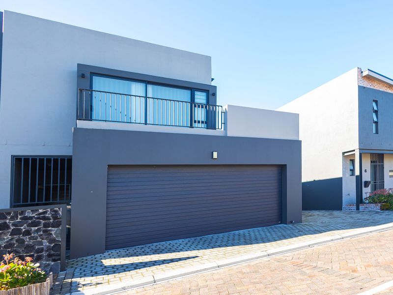 Modern double-story residential facade featuring a large automated garage door, paved driveway, and an upper-level balcony with a sleek metal balustrade.