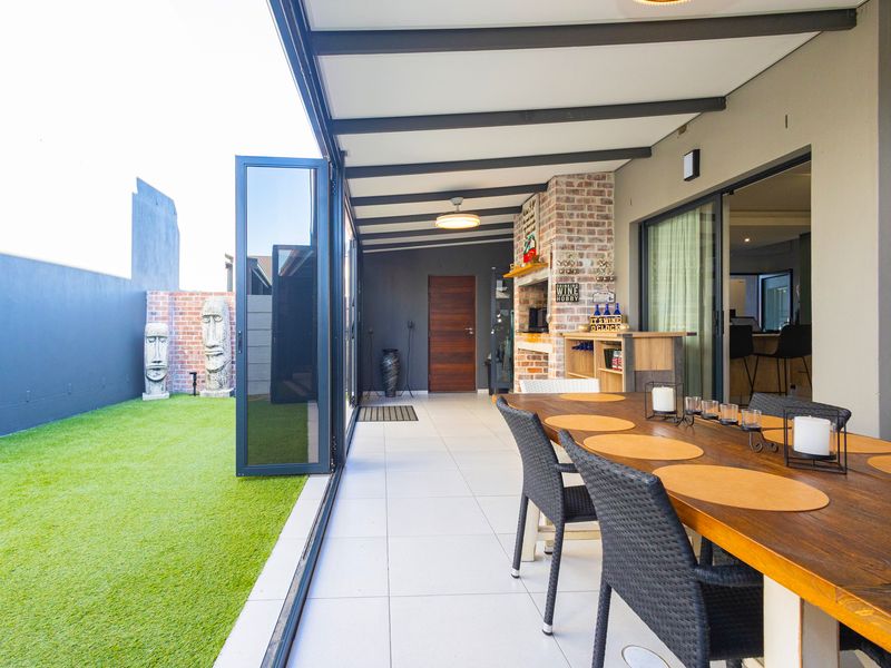 A spacious covered patio featuring a built-in braai, tiled flooring, and seamless stacking doors that open onto a neat artificial lawn area.