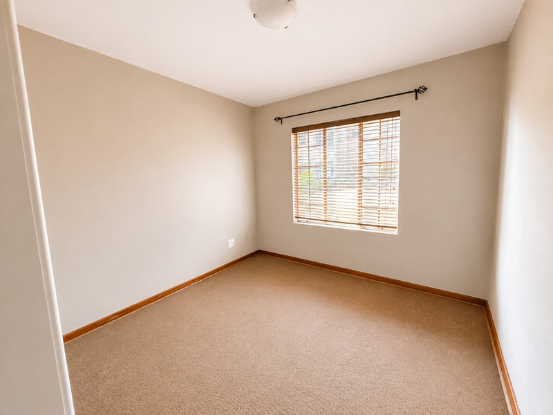 A clean, empty bedroom featuring neutral wall tones, carpeted flooring, and a window fitted with venetian blinds.