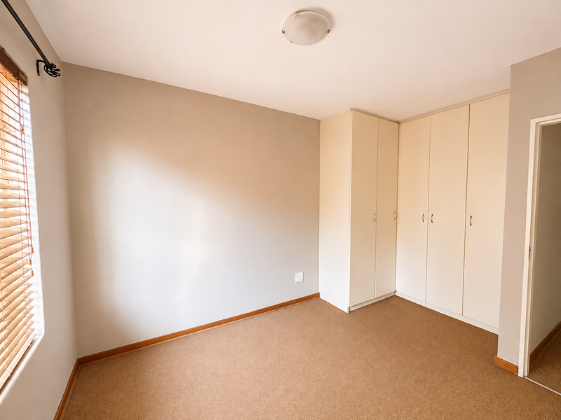 A clean, empty bedroom featuring neutral walls, carpeted flooring, built-in cupboards, and wooden blinds, offering a versatile space ready for personalization.