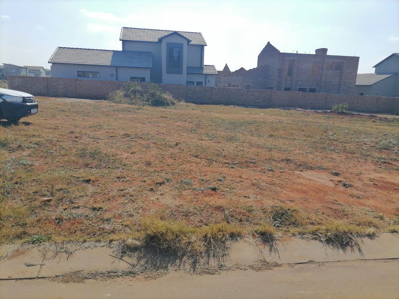 A vacant residential plot of land featuring a brick boundary wall and direct access to a paved road in a developing neighborhood.