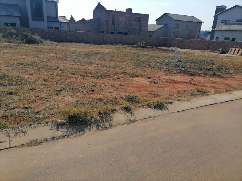 A vacant residential plot of land featuring a level surface, boundary wall, and direct access to a paved street in a developing estate.