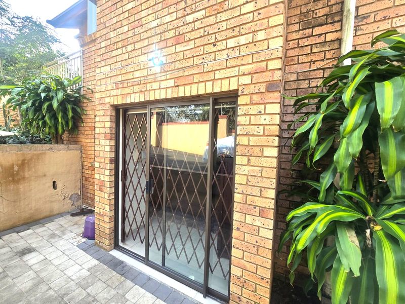 Rear exterior view of the apartment featuring a face brick facade, a sliding glass door with a security gate, and a paved patio area.