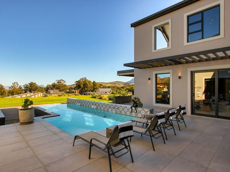 A modern outdoor pool area featuring a tiled patio, a decorative mosaic water feature wall, and a pergola, overlooking a landscaped garden and mountain views.