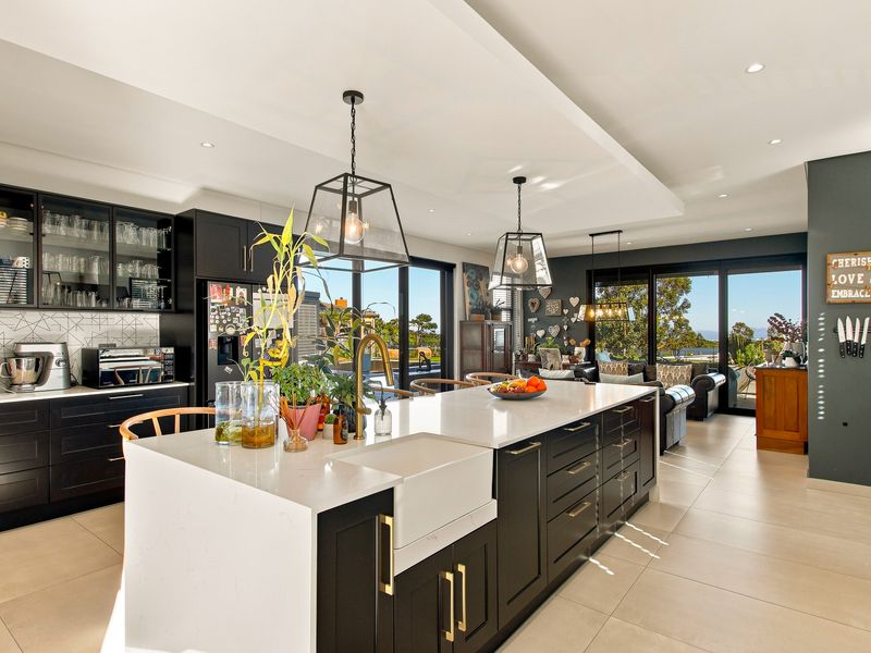 Modern open-plan kitchen featuring a large island with farmhouse sink, quartz countertops, dark cabinetry, and seamless flow to the living area with garden views.