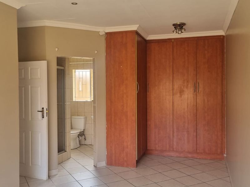 A spacious bedroom featuring built-in wooden cupboards, tiled flooring, and an attached en-suite bathroom with a shower.