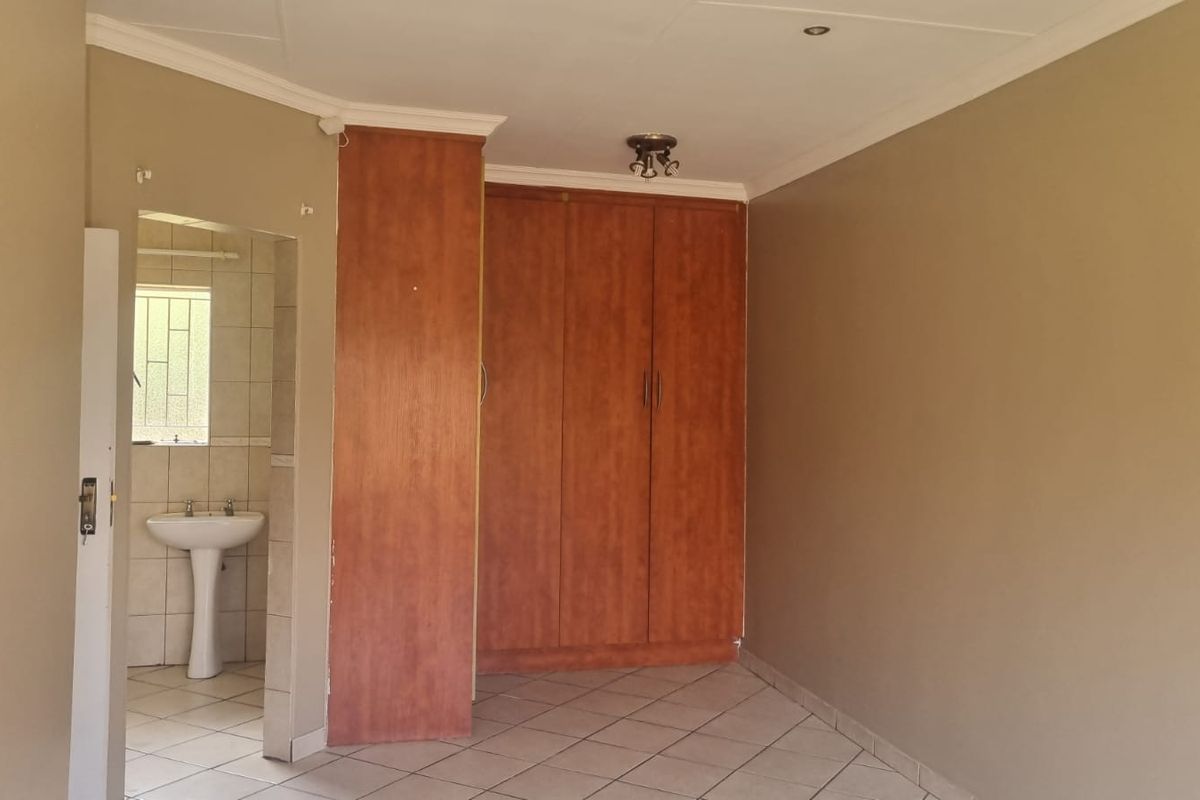 A spacious bedroom featuring built-in wooden cupboards, tiled flooring, and an attached en-suite bathroom with a pedestal basin.