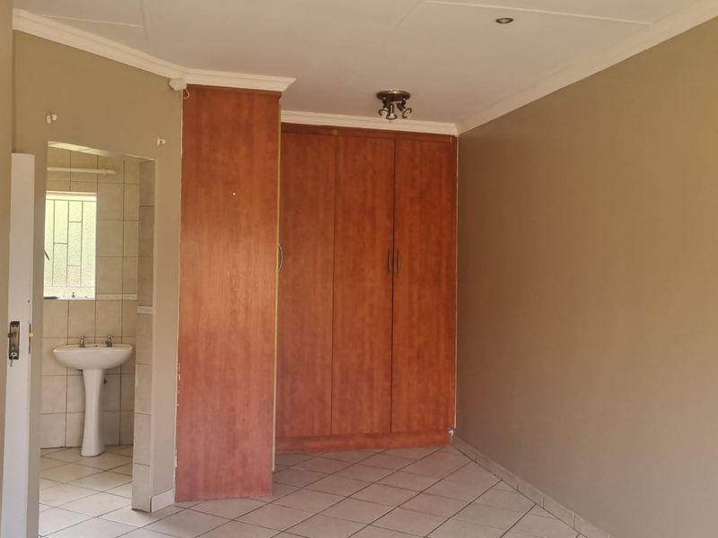 A spacious bedroom featuring built-in wooden cupboards, tiled flooring, and an attached en-suite bathroom with a pedestal basin.