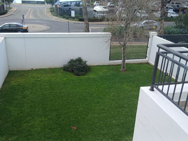 A private ground-level garden area featuring a manicured lawn, enclosed by white boundary walls and metal fencing, viewed from an elevated balcony.
