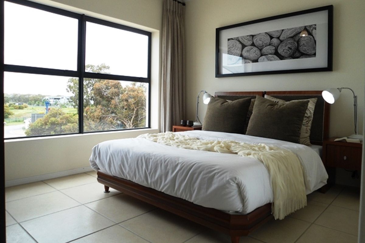 A well-lit bedroom featuring large windows with natural views, neutral tiled flooring, and clean wall finishes.