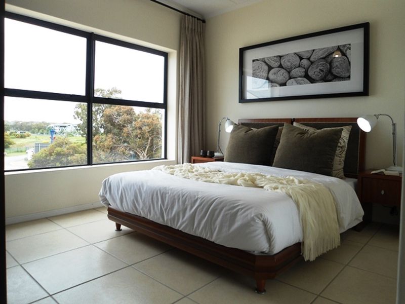 A well-lit bedroom featuring large windows with natural views, neutral tiled flooring, and clean wall finishes.