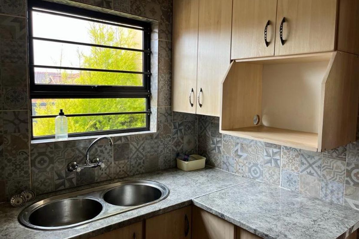 A functional scullery area featuring double stainless steel sinks, laminate countertops, ample built-in cabinetry, and decorative patterned wall tiling.
