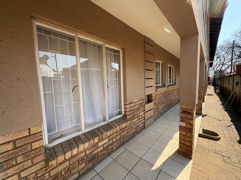 A covered outdoor patio area featuring tiled flooring, brick pillar supports, and security-barred windows along the building exterior.