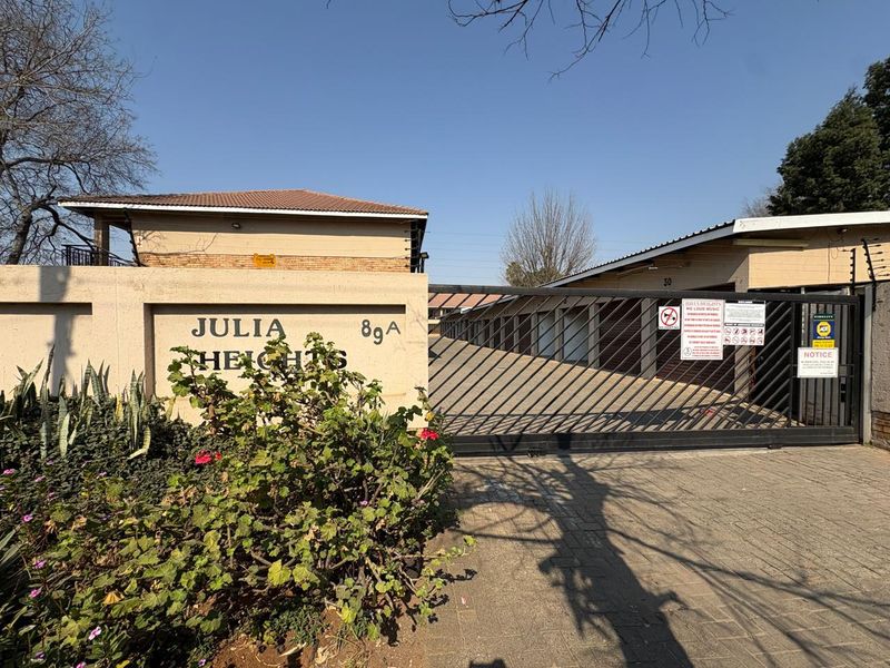 Front entrance view of Julia Heights apartment complex featuring a secure gated access point, paved driveway, and perimeter wall with electric fencing.