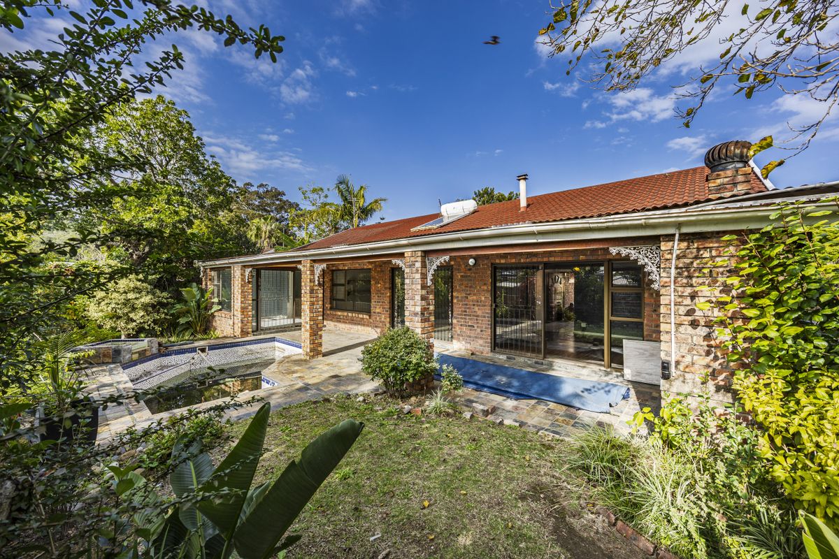 Rear view of a brick house featuring a covered patio, swimming pool, and established garden with mature trees.