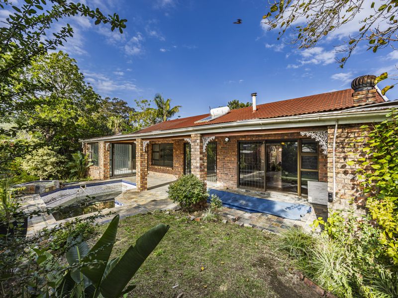 Rear view of a brick house featuring a covered patio, swimming pool, and established garden with mature trees.
