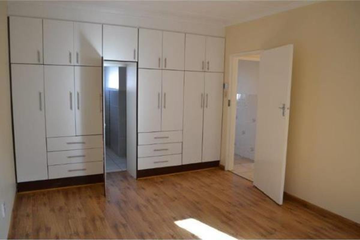 A clean, empty bedroom featuring extensive white built-in cupboards, laminate flooring, and direct access to an adjoining room.