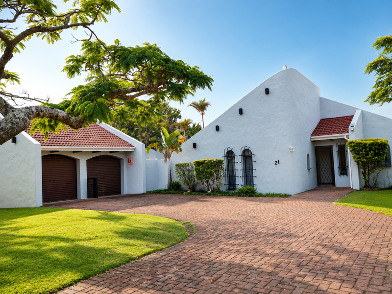 A well-maintained front elevation featuring a double garage, brick paved driveway, and a distinctive white facade with red tiled roofing.