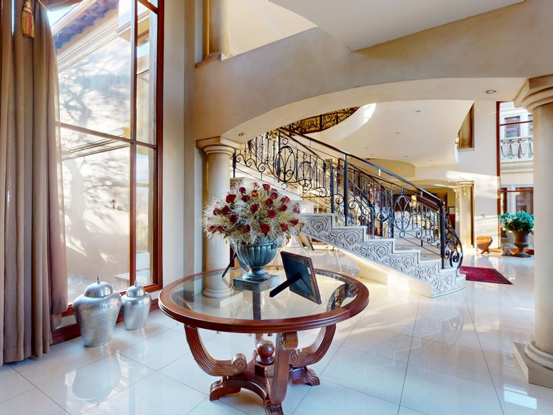 Grand entrance hall featuring high ceilings, floor-to-ceiling windows, polished tile flooring, and an elegant staircase with ornate wrought iron and stone detailing.
