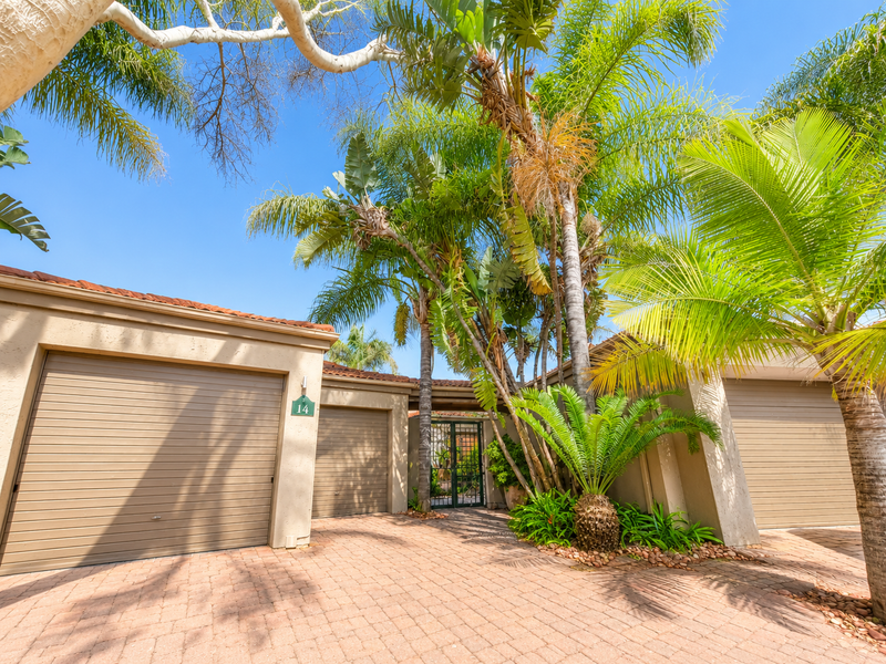 Front exterior view of the property featuring a paved driveway, double garage, and established garden with mature palm trees.