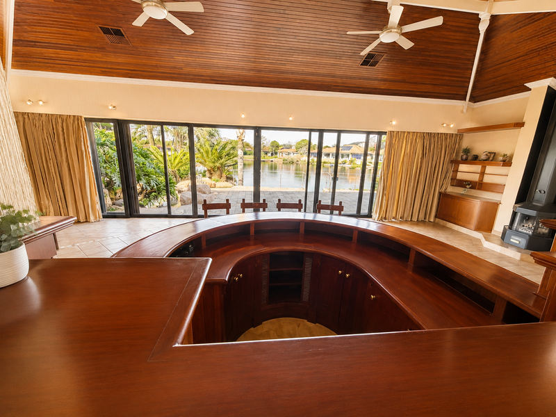 Spacious bar area featuring a large curved timber counter, vaulted timber ceiling, built-in fireplace, and expansive sliding doors opening to a waterfront view.