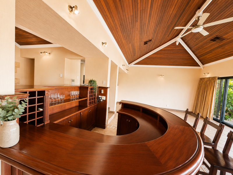 A well-appointed home bar featuring a curved wooden counter, custom cabinetry with integrated wine storage, and a vaulted timber ceiling with a ceiling fan.