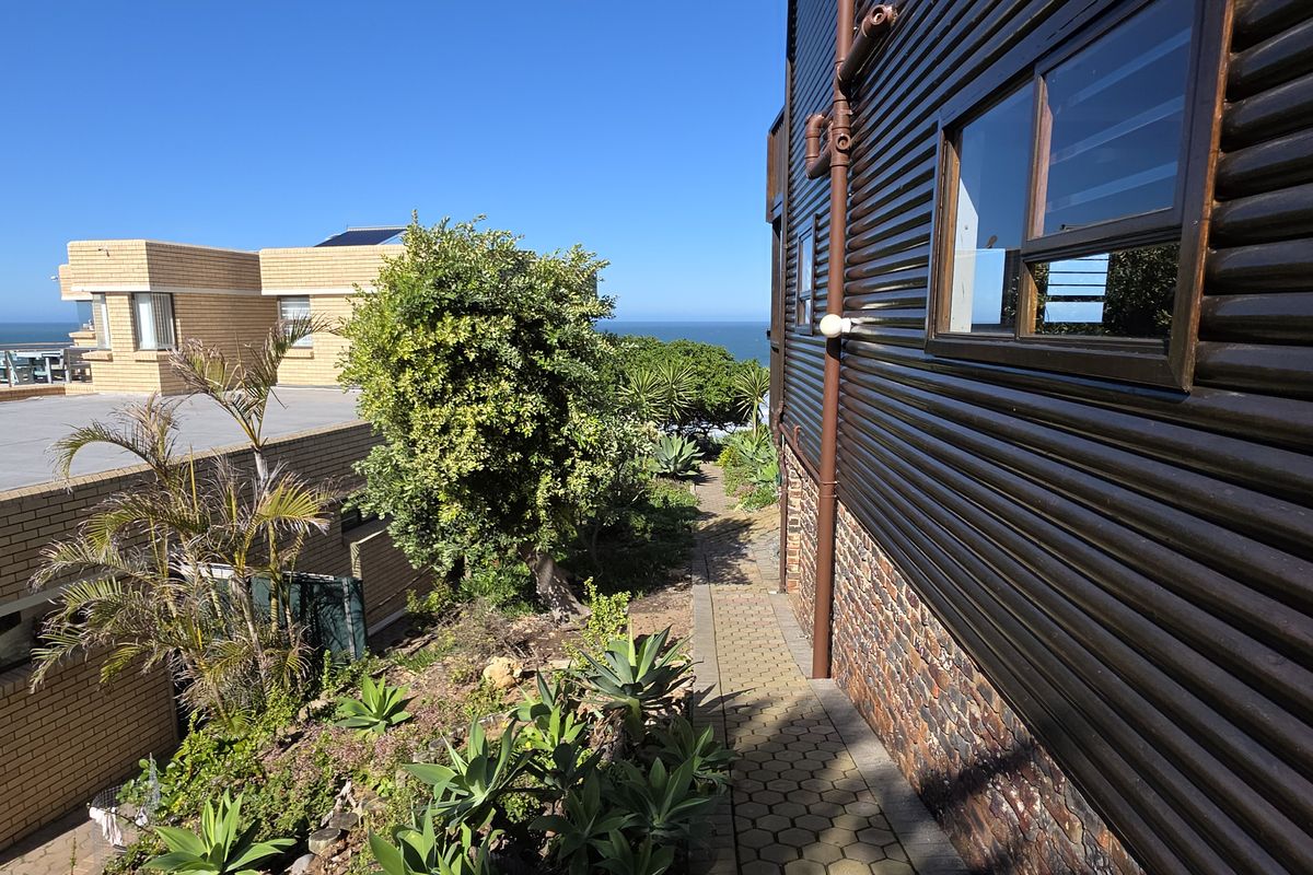 Side view of the property showing corrugated metal cladding, stone masonry base, and a paved walkway leading towards the rear garden and ocean view.