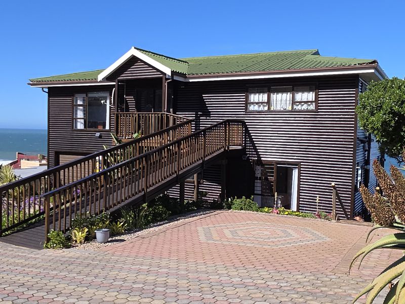 A well-maintained coastal home featuring dark corrugated metal cladding, a green roof, and a timber ramp entrance, set against a scenic ocean backdrop.