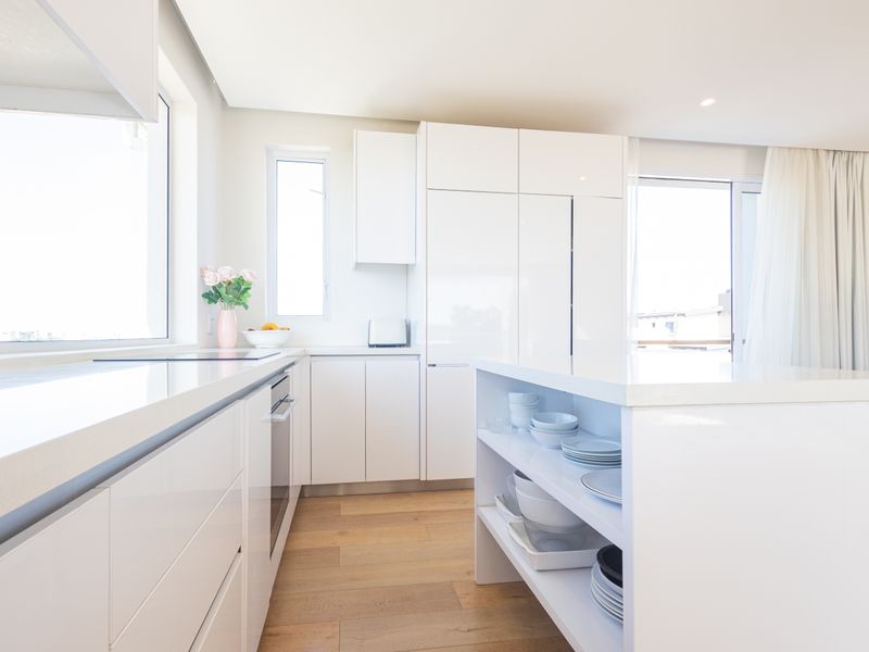 Modern, bright kitchen featuring sleek white cabinetry, stone countertops, an integrated oven, and a functional island with open shelving, set on hardwood flooring.