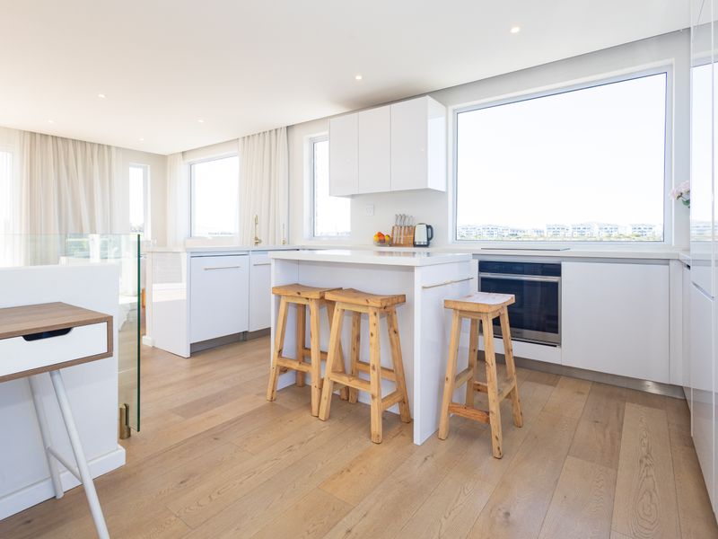 Modern, bright kitchen featuring white cabinetry, engineered stone countertops, a central island, and high-quality hardwood flooring with large windows.