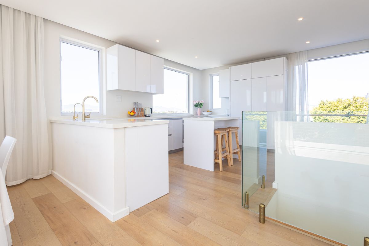 Modern, bright kitchen featuring white gloss cabinetry, stone countertops, brass fixtures, and light timber flooring, seamlessly integrated with an open-plan living area.