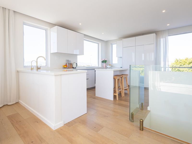 Modern, bright kitchen featuring white gloss cabinetry, stone countertops, brass fixtures, and light timber flooring, seamlessly integrated with an open-plan living area.