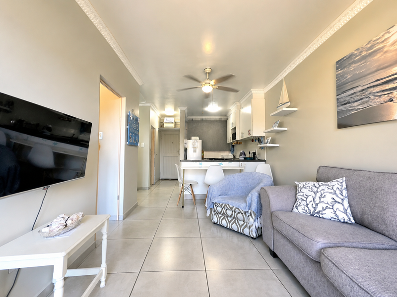 Open-plan living area featuring tiled flooring, crown molding, a ceiling fan, and a compact kitchen with built-in cabinetry.