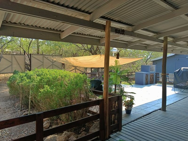Covered outdoor patio area featuring a built-in braai, timber decking, and a corrugated metal roof, overlooking a garden space.
