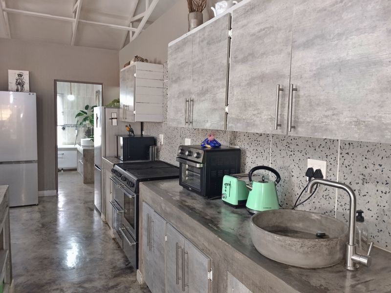 A modern kitchen featuring concrete countertops, a unique concrete basin, grey wood-look cabinetry, and exposed ceiling rafters, leading into an adjacent room.
