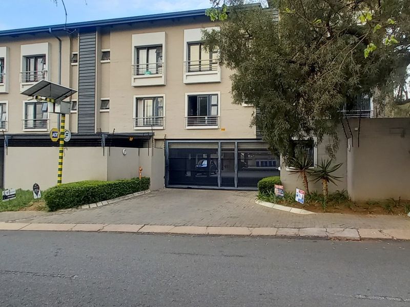 Front elevation of a modern apartment complex featuring a paved driveway, secure automated gate access, covered parking, and balconies on upper levels.