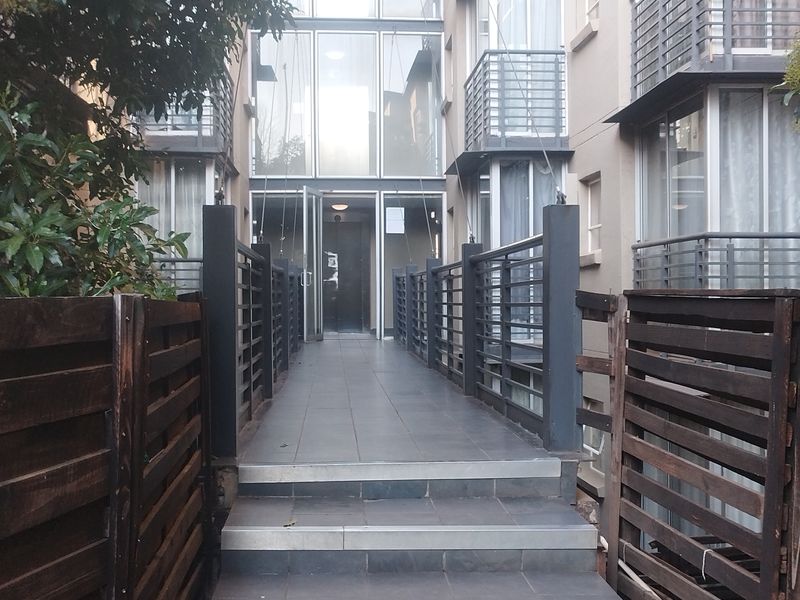 Exterior view of an apartment building entrance featuring a tiled walkway, metal railings, and a modern glass facade with timber privacy screens.