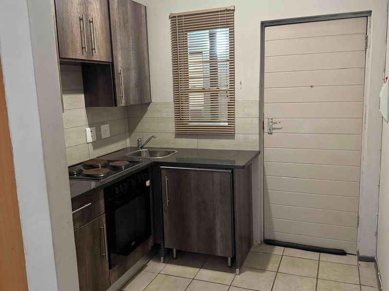 Compact kitchen featuring dark wood-look cabinetry, built-in oven and hob, stainless steel sink, and tiled flooring with a neutral backsplash.