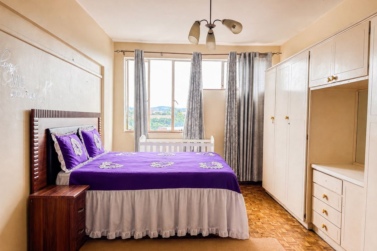 A bedroom featuring parquet flooring, a large built-in wardrobe with integrated drawers, and natural light from a window.