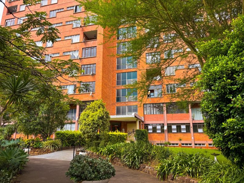 A view of the front facade of a multi-story brick apartment building surrounded by mature trees and landscaped garden areas.