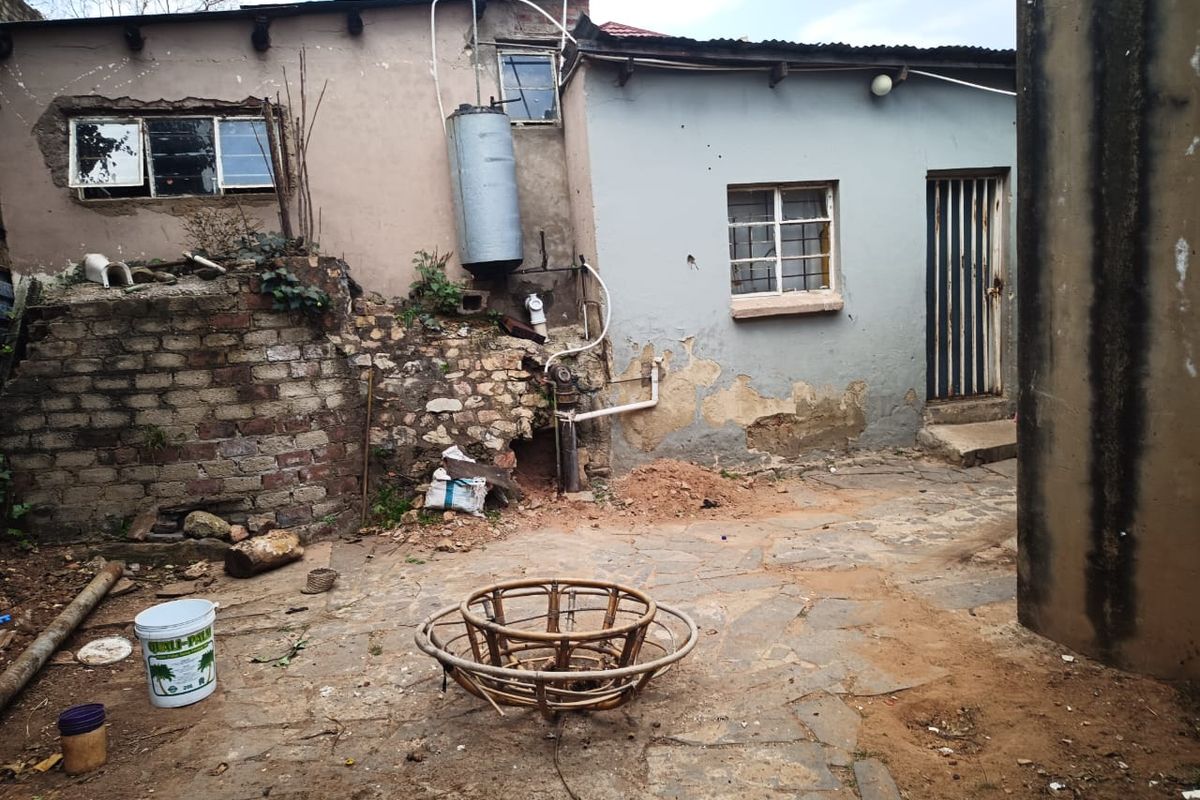 Rear courtyard area featuring stone paving, exposed exterior plumbing, and an electric geyser, showing significant signs of wear and maintenance requirements.
