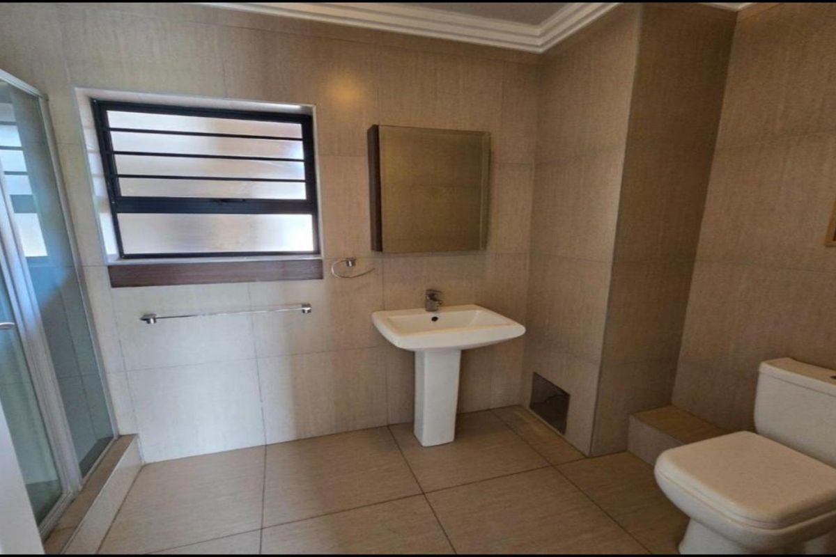 A functional bathroom featuring a pedestal basin, shower enclosure, toilet, and neutral wall and floor tiling with a louvered window for ventilation.