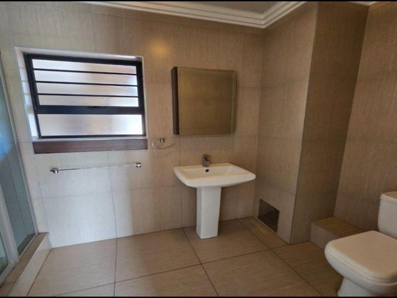 A functional bathroom featuring a pedestal basin, shower enclosure, toilet, and neutral wall and floor tiling with a louvered window for ventilation.