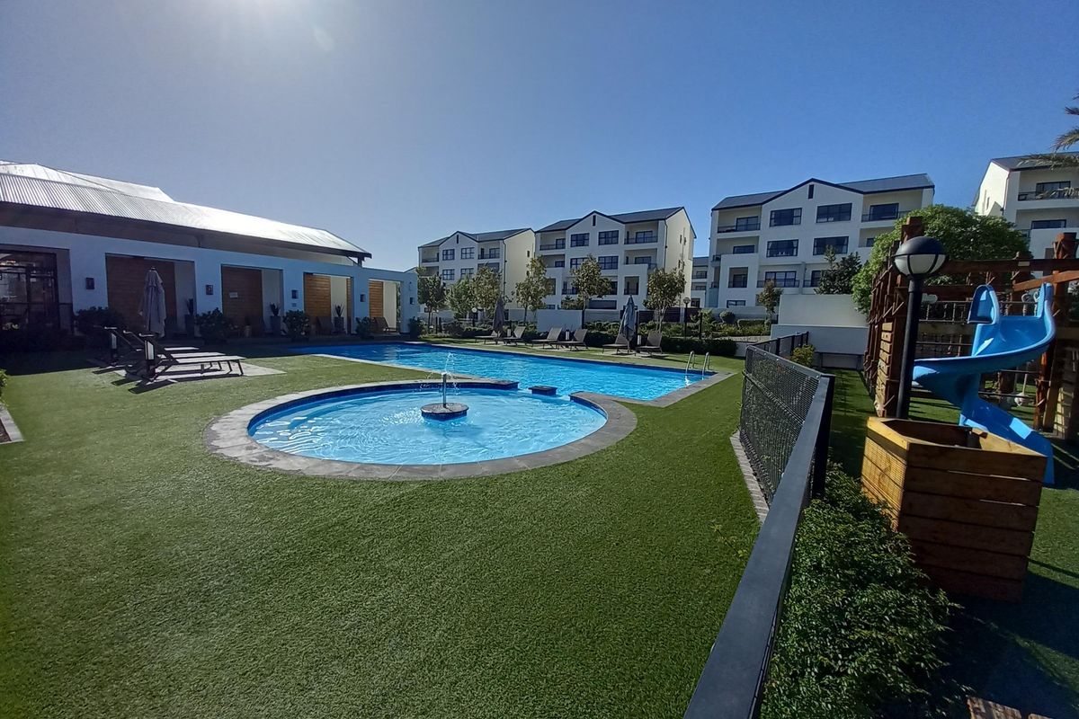 A well-maintained communal swimming pool area featuring a fountain, artificial lawn, and surrounding apartment complex buildings.