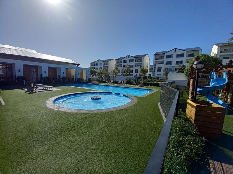A well-maintained communal swimming pool area featuring a fountain, artificial lawn, and surrounding apartment complex buildings.