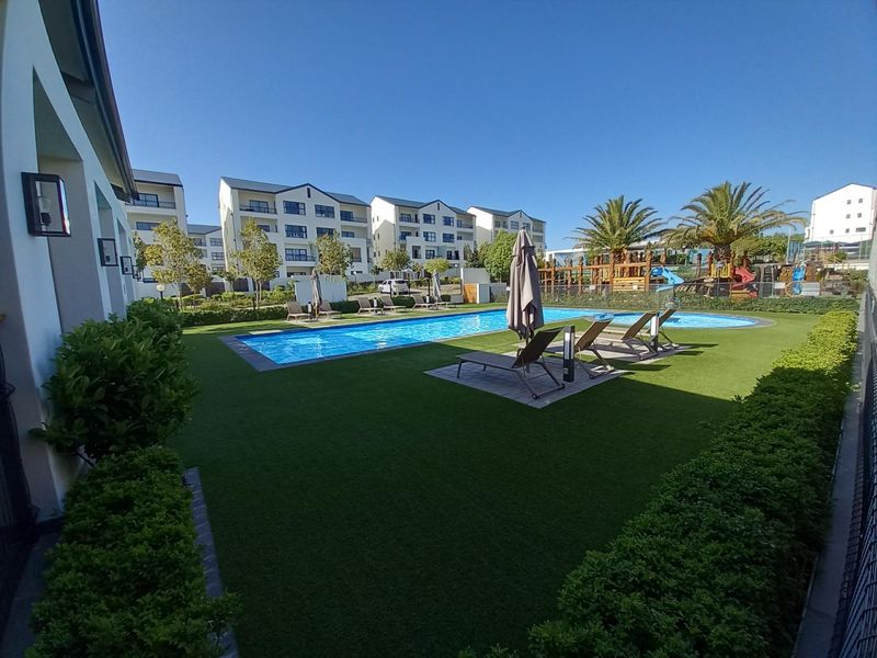 A well-maintained communal swimming pool area featuring artificial turf, landscaped hedging, and modern exterior lighting within a residential apartment complex.