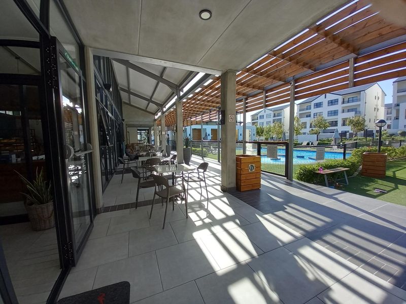 A spacious covered patio area featuring tiled flooring and a timber pergola, overlooking the communal swimming pool and landscaped garden area.